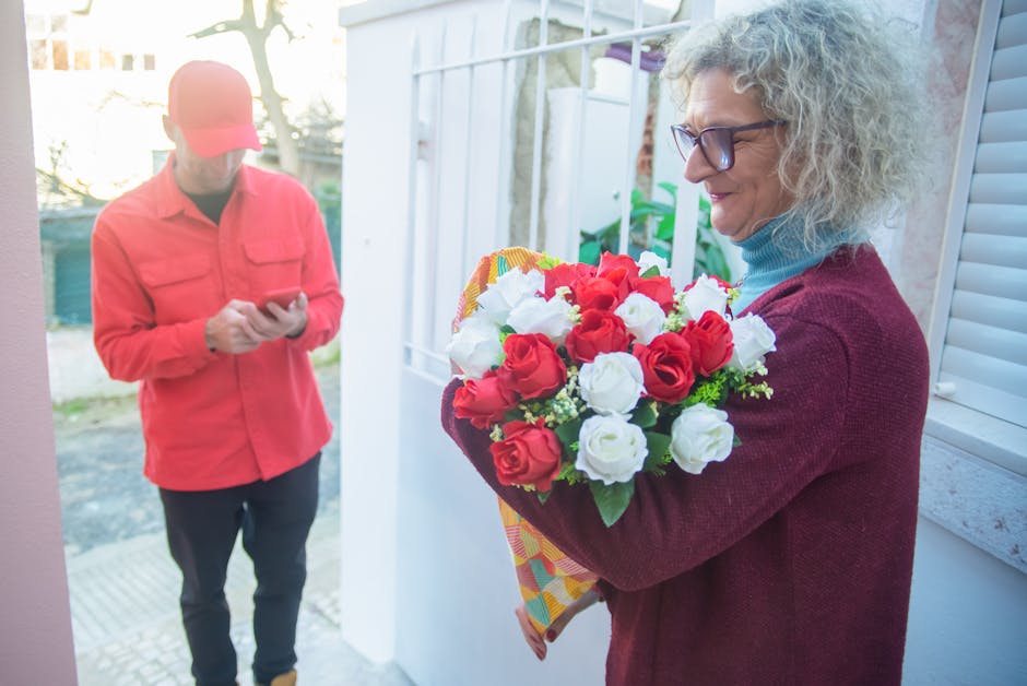 A woman with curly gray hair, wearing glasses, a blue turtleneck, and a burgundy sweater, holds a colorful bouquet of fresh red and white roses wrapped in floral paper, standing at her front door duri