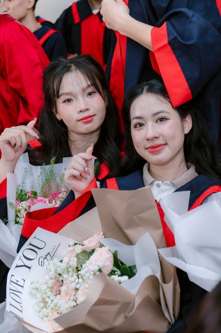 A close-up of two young women dressed in graduation gowns with black and red accents, holding and surrounded by colorful floral bouquets including pink and white flowers with green foliage, wrapped in