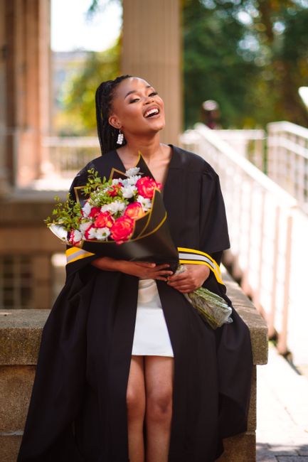 A young woman dressed in a black graduation gown with yellow trim is seated on an outdoor stone step, holding a large, vibrant bouquet of fresh flowers featuring pink roses, white daisies, and green f