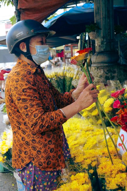 A woman wearing a black helmet and a blue face mask arranges a small bouquet of yellow and red roses at an outdoor flower market. She is dressed in an orange jacket with a black pattern and a colorful