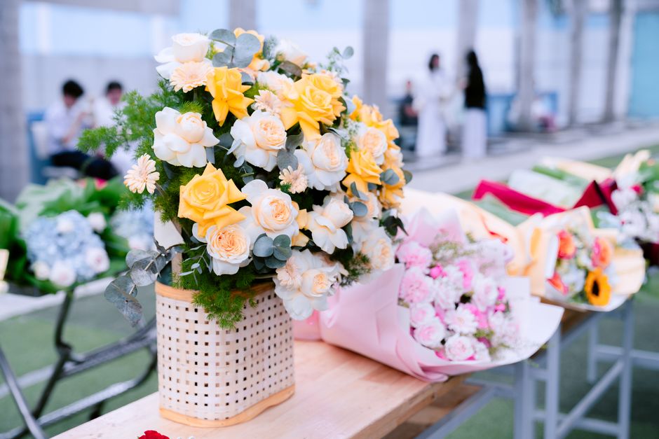 A floral arrangement featuring a bouquet of yellow and white roses, along with white garden flowers, placed in a woven white basket with green foliage accents. The bouquet is positioned on a wooden ta