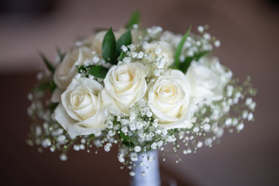 A floral bouquet featuring creamy white roses with softly curled petals, surrounded by delicate white baby's breath flowers and a few green leaves, arranged in a compact, rounded style. The bouquet is
