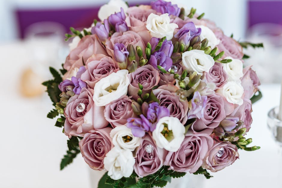 A close-up image of a floral bouquet featuring large, soft pink roses, white lisianthus, and small purple freesia, arranged in a rounded, compact style. The bouquet is accented with green foliage and 