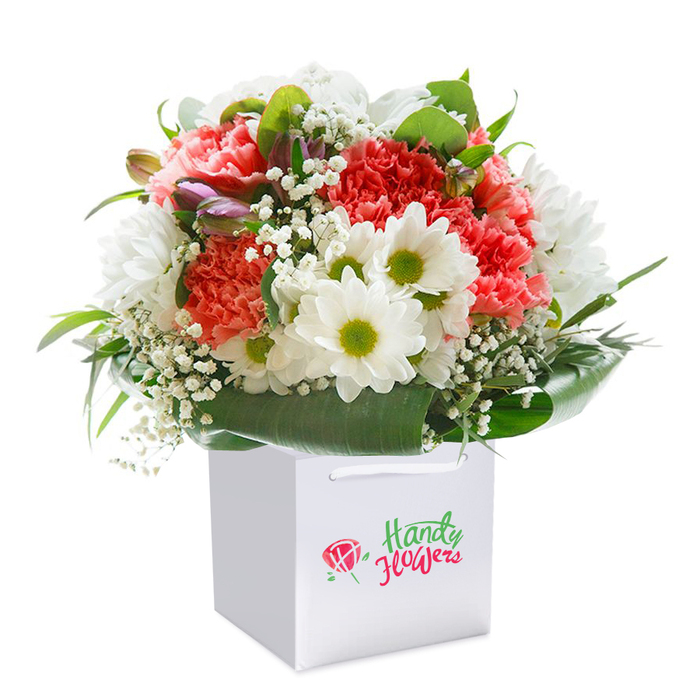 Presented upright in a crisp white gift bag against a flawless white background, this beautifully designed bouquet captures the charm and freshness characteristic of passionate floristry in Luton. Plush coral-pink carnations, distinguished by their intricate, ruffled petals, cluster vibrantly together, accented by the lively presence of bright white daisy-like chrysanthemums whose lime-hued centres create visual harmony. Dispersed amidst these are fluttering sprays of white baby's breath, gently softening the display and lending a gentle, ethereal air. Subtle touches of purple-tinged buds or closed flowers peep from the left, hinting at new growth and tender moments yet to come. The bouquet is gracefully encircled by lush, glossy greenery and deep, broad leaves, which wrap the base in a protective embrace reminiscent of Luton's lovingly tended gardens. With its purposeful rounded silhouette, the bouquet conveys an inviting abundance-perfect for celebrating birthdays, anniversaries, or brightening the day of a loved one nearby. Sturdy ribbon handles atop the white gift bag add a thoughtful convenience, echoing the modern ease of arranging same-day floral delivery within Luton's vibrant urban landscape. Every detail, from the careful choice of blooms to the precise arrangement, reflects the artistry and pride of a trusted local provider, ensuring a memorable and heartfelt experience.