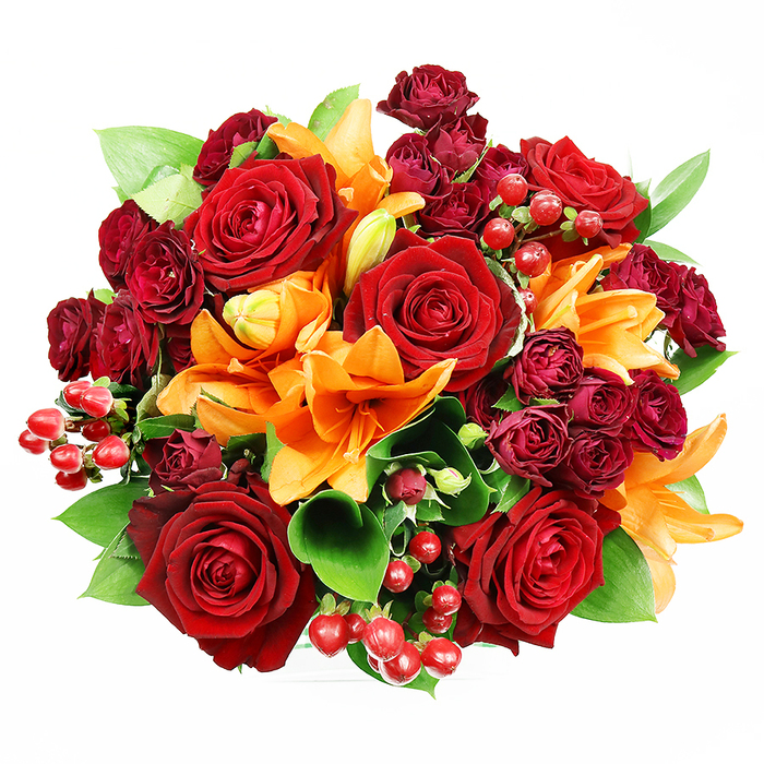 From a top-down perspective against spotless white, this richly composed bouquet reads like a jewel box opened on a table, each element chosen for colour, texture and emotive weight. Dominating the scene are sumptuous, fully opened deep red roses, their layered petals soft to the eye and evocative of classic romance; they anchor the design with a warm, velvety gravity. Interspersed are flamboyant orange Asiatic lilies whose trumpet forms and visible stamens introduce a brighter, optimistic energy - some display dark speckles in their throats, while others linger as tight buds, promising sequential blooming and a sense of unfolding. Smaller, concentrated clusters of burgundy spray roses add a velveteen depth, their miniature blooms packing colour into the negative spaces and creating luxurious contrast with the larger roses. Glossy hypericum berries, the colour of ripe cranberries, are scattered through the arrangement, catching highlights and offering a tactile, berry-like punctuation against the petals. Generous, glossy green foliage-suggestive of ruscus or salal-threads through the design, its fresh, clean leaves cooling the palette and providing structural rhythm that leads the eye across the bouquet. The lighting is gentle and even, chosen to emphasize petal surface, subtle shadowing and the bouquet's three-dimensional fullness. I picture this arrangement on a Luton dining table after a walk through Wardown Park in crisp air, or sent to celebrate a milestone in Stopsley or Bury Park; it's equally suited to anniversaries, heartfelt thank-yous, or an autumnal centrepiece. The overall mood is opulent yet approachable, a carefully composed expression of warmth and celebration by experienced hands.